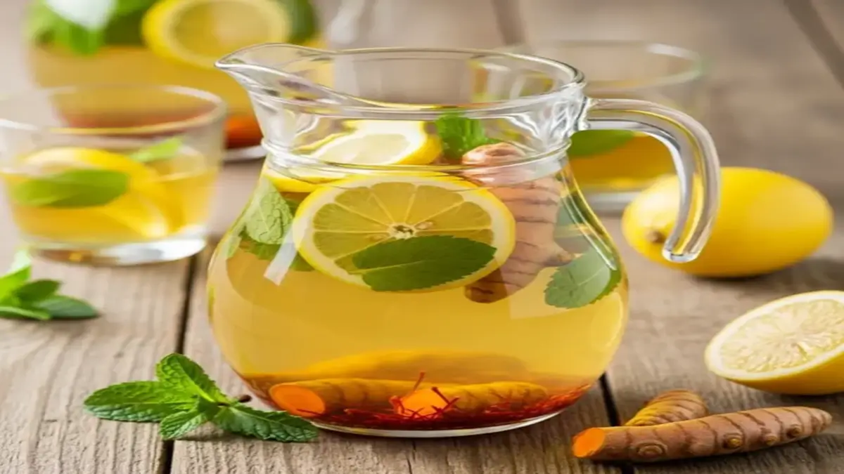 Copo de vidro com água de açafrão dourada, rodelas de limão fresco, raiz de cúrcuma fresca e folhas de hortelã sobre fundo de madeira rústica.