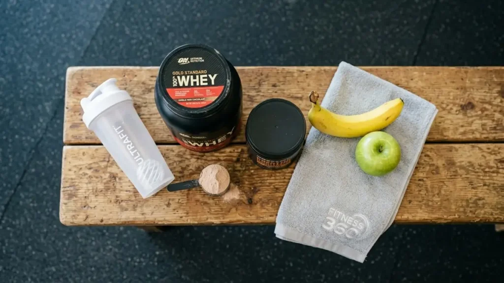Sports Supplements - Flat lay of whey protein, creatine, shaker, and fresh fruits on a gym bench.
